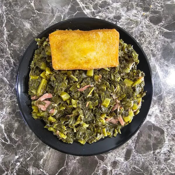 A black plate of collard greens with ham, topped with a golden-brown cornbread square.